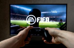 Mãos segurando controle de videogame. TV exibe jogo de futebol "EA SPORTS FIFA" em estádio.