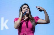 Mulher de vestido rosa discursa, segurando microfone e gesticulando. Fundo azul com parte de um 'K' branco.