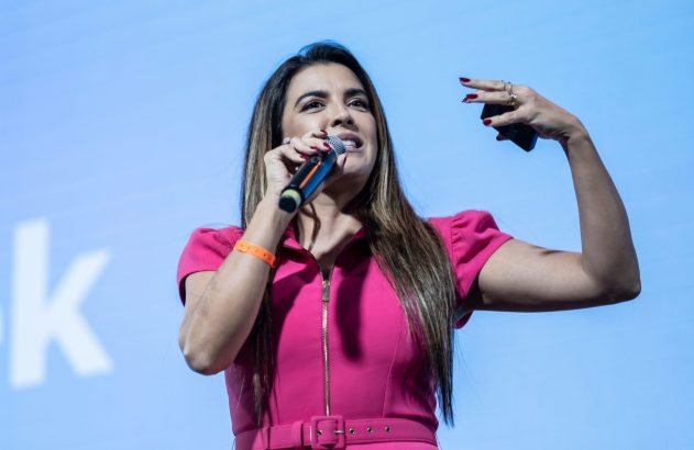 Mulher de vestido rosa discursa, segurando microfone e gesticulando. Fundo azul com parte de um 'K' branco.