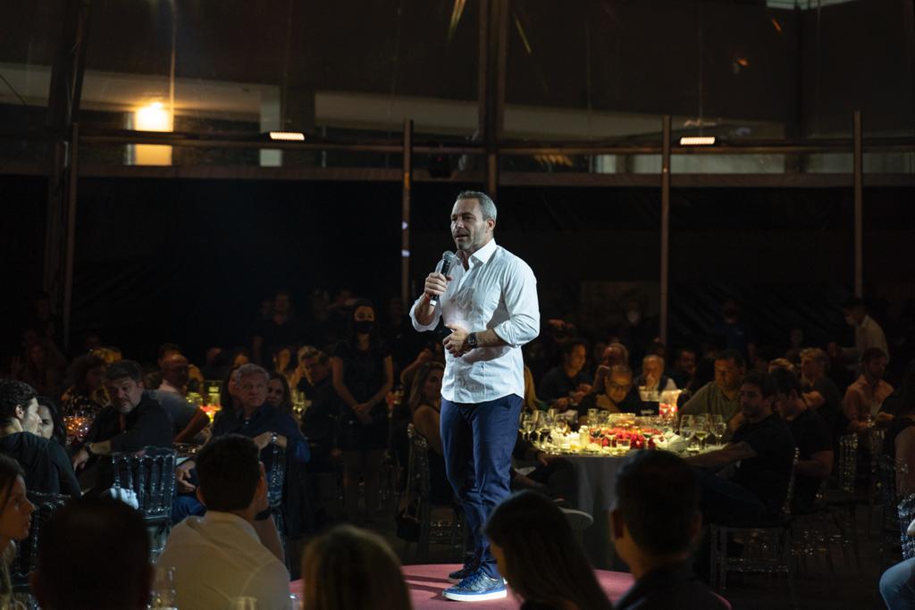 Palestrante masculino em palco escuro, falando para público sentado em mesas em evento.