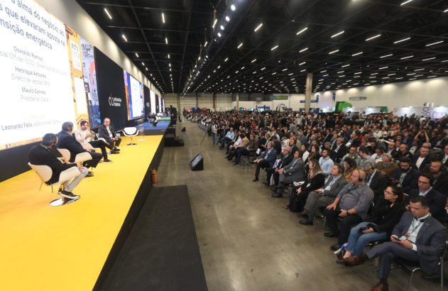 Painel de debate automotivo com palestrantes em palco amarelo e numerosa plateia em salão de eventos.