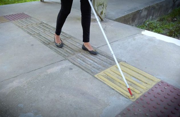 Pessoa com bengala branca navegando em calçada com piso tátil direcional (amarelo) e de alerta (vermelho).