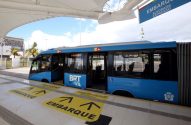 Ônibus articulado azul BRT na estação, portas abertas. Setas amarelas "Embarque" no chão. Placa superior "Alvorada Expresso".
