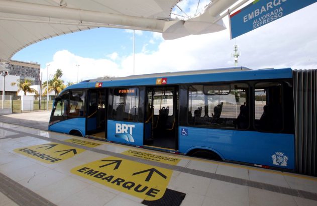 Ônibus articulado azul BRT na estação, portas abertas. Setas amarelas "Embarque" no chão. Placa superior "Alvorada Expresso".
