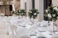 Recepção de evento com mesas e cadeiras brancas, arranjos florais de folhagens e flores brancas, em salão elegante.