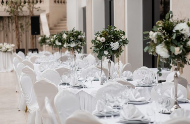 Recepção de evento com mesas e cadeiras brancas, arranjos florais de folhagens e flores brancas, em salão elegante.