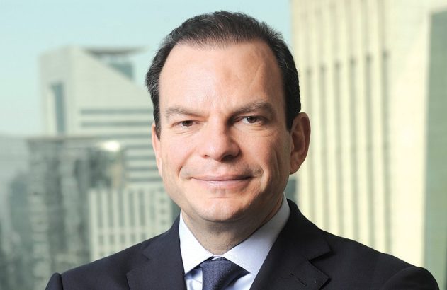 Homem de terno sorrindo para a câmera, com prédios desfocados ao fundo.