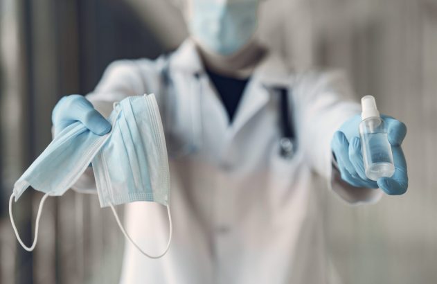 Profissional de saúde com máscara e luvas oferece máscaras e spray desinfetante.