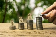 Mão adiciona moeda a pilha de dinheiro em crescimento. Quatro pilhas de moedas com sacos de dólar em mesa de madeira.