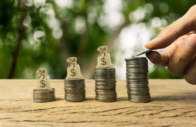 Mão adiciona moeda a pilha de dinheiro em crescimento. Quatro pilhas de moedas com sacos de dólar em mesa de madeira.