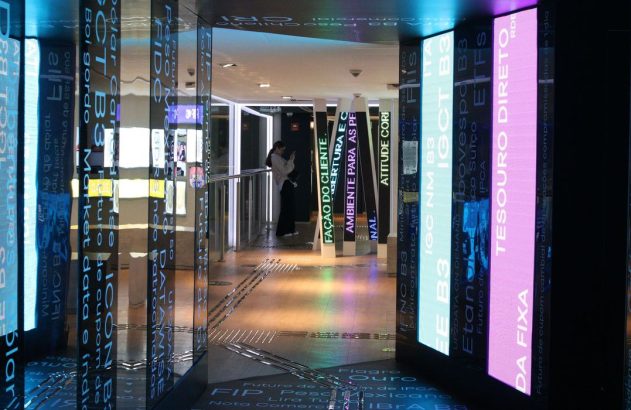 Hall com pilares digitais coloridos exibindo termos financeiros e uma pessoa olhando o celular ao fundo.