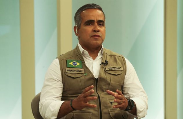 Ronaldo Bento, Ministro da Cidadania, em colete com bandeira do Brasil, gesticula enquanto fala.