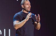 Homem com barba e cabelos grisalhos, camiseta preta e calça roxa, gesticula em palco com letreiro "UM" e "SIL" ao fundo.