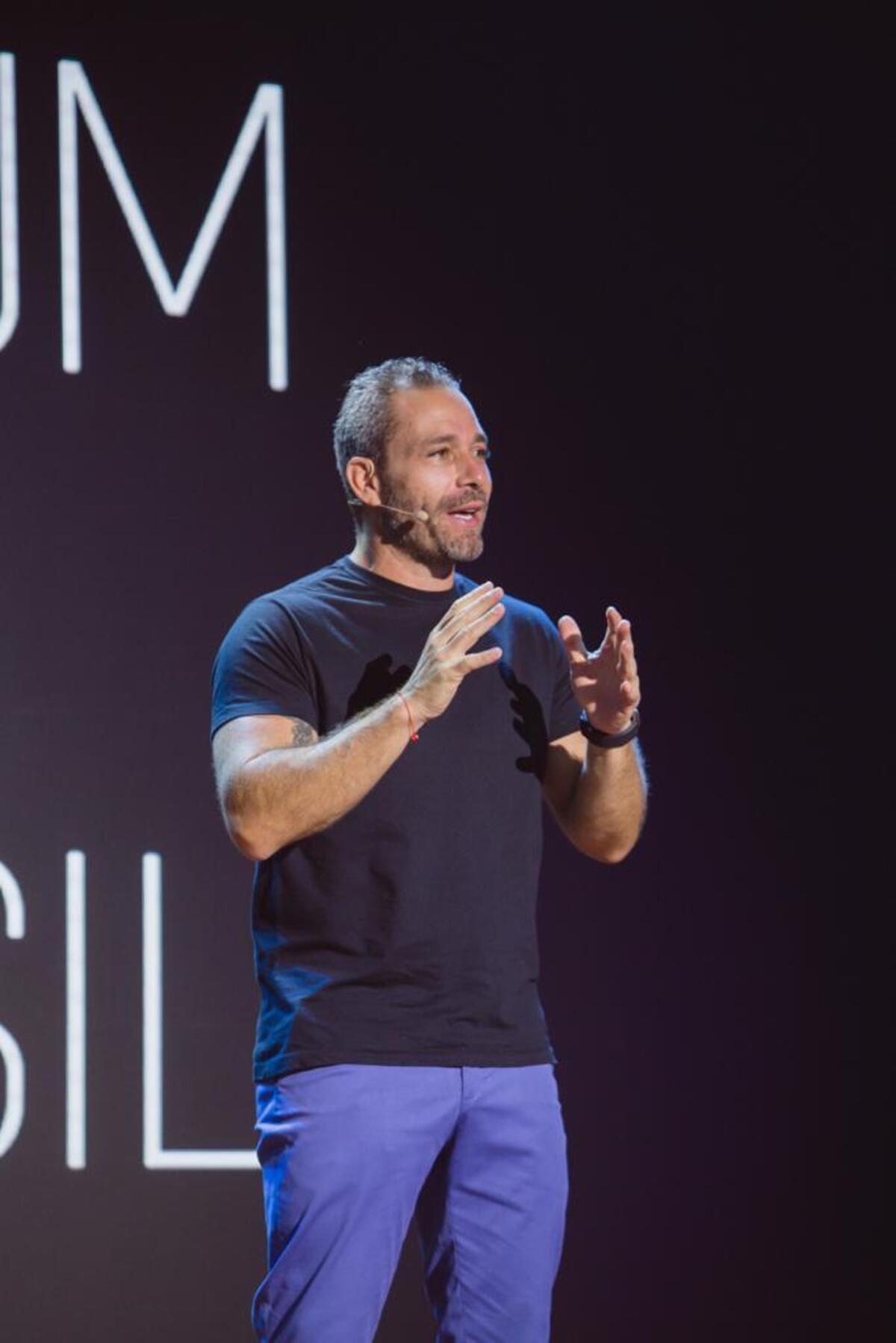 Homem com barba e cabelos grisalhos, camiseta preta e calça roxa, gesticula em palco com letreiro "UM" e "SIL" ao fundo.