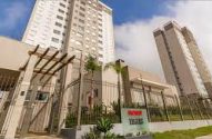 Entrada de condomínio residencial com duas torres de apartamentos, portaria e paisagismo. Dia claro.