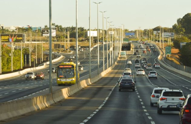 Rodovia de múltiplas pistas com tráfego intenso de veículos e um ônibus amarelo à esquerda.