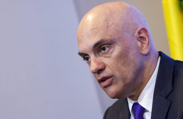 Homem careca de terno e gravata roxa, boca entreaberta, olhando para baixo.