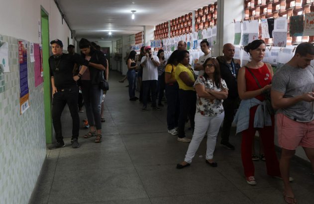 Pessoas em fila aguardando em corredor interno com paredes de azulejo e trabalhos expostos.