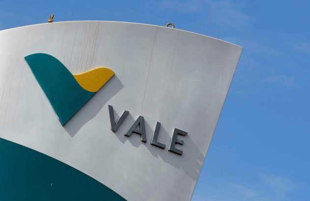 Logotipo da Vale: símbolo em azul-petróleo e amarelo, com o nome 'VALE' em cinza sobre superfície branca curva e céu azul.