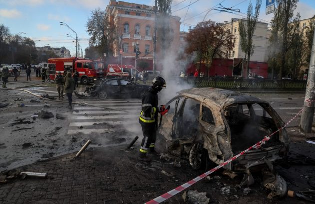 Bombeiro em rua com fumaça e escombros, apagando carro queimado. Outro carro e caminhões de bombeiro no fundo.