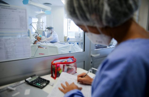 Profissionais de saúde em hospital: um escreve e outro assiste paciente em leito, separados por vidro.