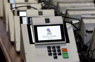 Várias urnas eletrônicas brasileiras enfileiradas. Uma mostra a tela inicial da Justiça Eleitoral e seu teclado de voto.