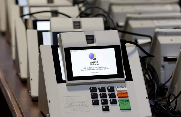 Várias urnas eletrônicas brasileiras enfileiradas. Uma mostra a tela inicial da Justiça Eleitoral e seu teclado de voto.