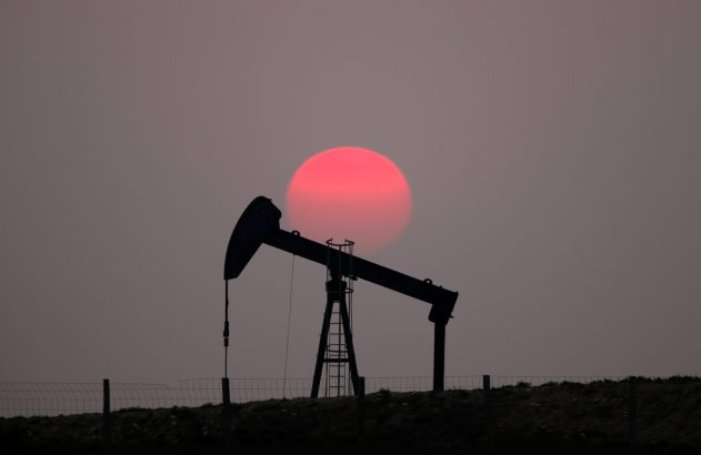 Silhueta de uma bomba de petróleo contra um sol vermelho intenso no crepúsculo. Uma cerca aparece no primeiro plano.