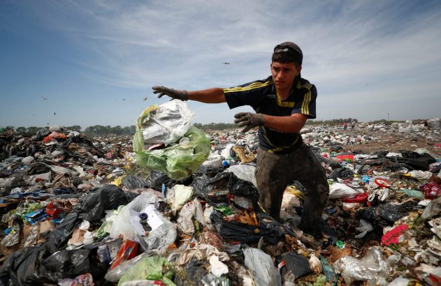 Jovem em lixão, lançando um saco de resíduos em meio a montanhas de lixo, sob céu parcialmente nublado.
