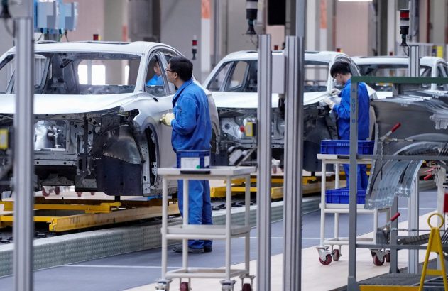 Trabalhadores em macacões azuis montam carrocerias em linha de montagem automotiva.