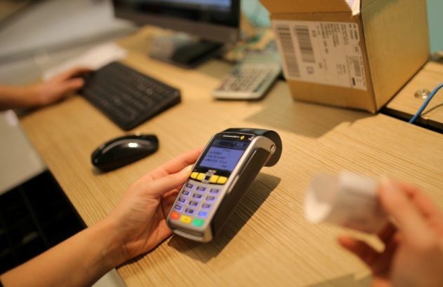 Mãos usando um terminal de pagamento eletrônico (POS) em uma mesa de trabalho. Fundo com teclado, mouse e pacote.
