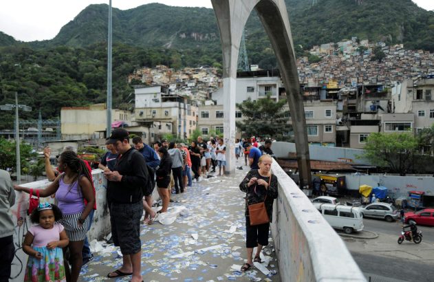 Pessoas em ponte com chão coberto de papéis. Favelas e montanhas ao fundo sob grande arco de concreto.