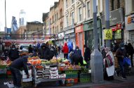 Pessoas com máscaras em mercado de rua com barracas de frutas e legumes. Prédios urbanos e arranha-céus ao fundo.