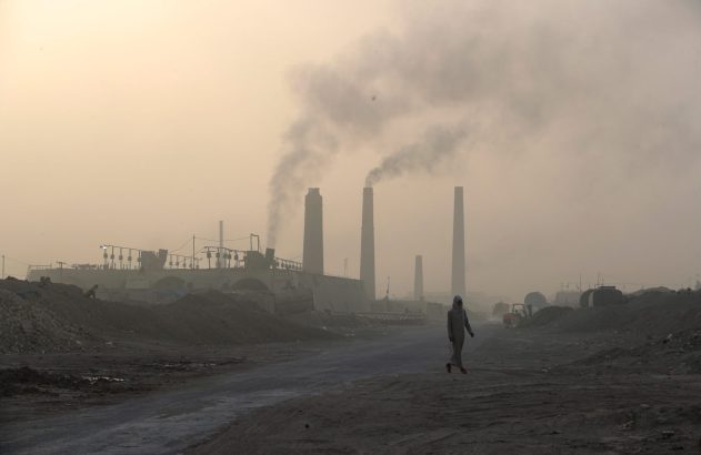 Pessoa anda em área industrial. Chaminés emitindo fumaça densa no céu cinzento, sobre chão árido e empoeirado.