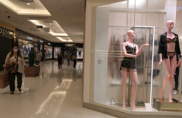 Mulher de máscara caminha em shopping. Vitrine com manequins de lingerie à direita. Pessoas ao fundo.