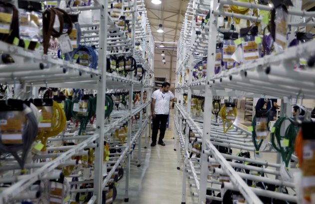Homem em camisa branca inspeciona prateleiras repletas de cabos coloridos em um armazém industrial.
