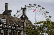 London Eye, bandeira britânica e prédio com chaminés sob céu cinzento. Avião e folhagem.