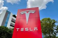 Letreiro vermelho da Tesla com logo branco, prédio moderno, árvores verdes e céu azul com nuvens.