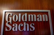 Letreiro "Goldman Sachs" com letras cinzas e moldura sobre fundo de madeira escura.