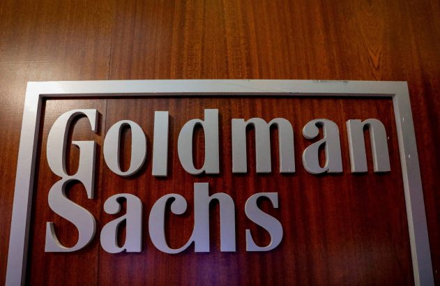 Letreiro "Goldman Sachs" com letras cinzas e moldura sobre fundo de madeira escura.