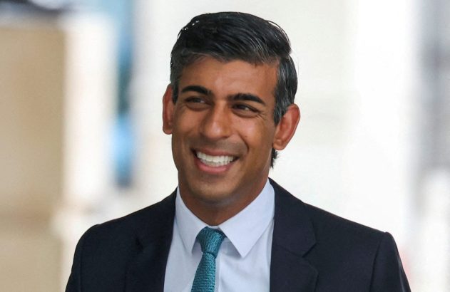 Close-up de Rishi Sunak sorrindo, com terno escuro e gravata azul-esverdeada.