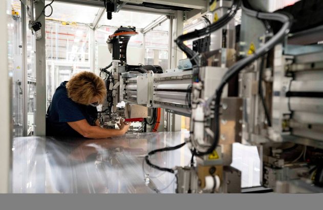 Mulher inclinada trabalha em máquina industrial, manipulando peças com ferramenta vermelha.