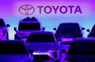 Múltiplos carros modernos da Toyota em exposição sob o logo da marca, com iluminação vibrante roxa e azul.