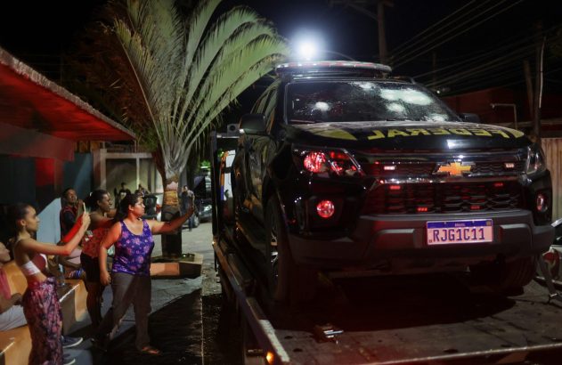 Viatura da Polícia Federal com para-brisa baleado no guincho, com pessoas observando à noite.