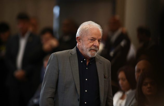 Lula, de blazer cinza e camisa azul, em evento interno, olhando para o lado.