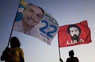 Pessoas silhuetadas seguram bandeiras de Bolsonaro (à esq.) e Lula (à dir.) contra o céu claro.