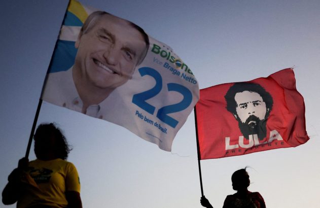 Pessoas silhuetadas seguram bandeiras de Bolsonaro (à esq.) e Lula (à dir.) contra o céu claro.