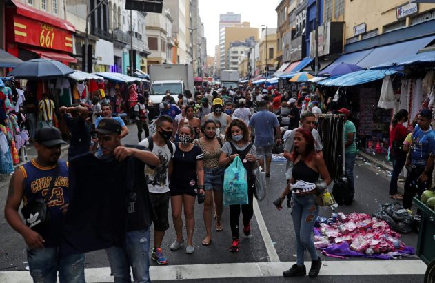 Rua de comércio movimentada com pedestres e barracas. Muitos usam máscaras.