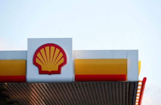 Logo da Shell (concha amarela e vermelha) no topo de um posto de combustível, com o céu claro ao fundo.