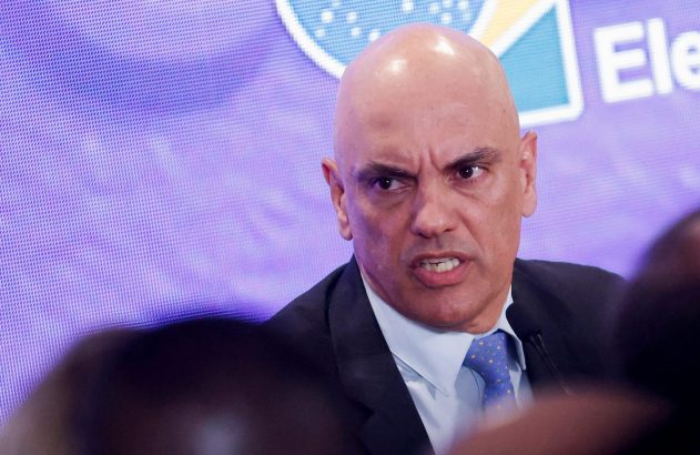 Homem careca de terno, com rosto franzido e boca aberta, em discurso. Fundo roxo pixelado.